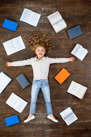 Happy child. Top view creative photo of little girl on vintage brown wooden floor. Girl lying near books, looking at camera and smilingの写真素材