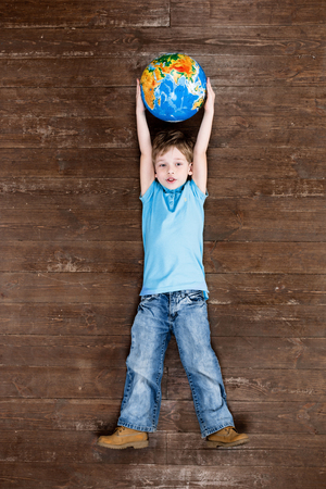 Happy child. Top view creative photo of little boy on vintage brown wooden floor. Boy holding globe, looking at camera and smilingの写真素材