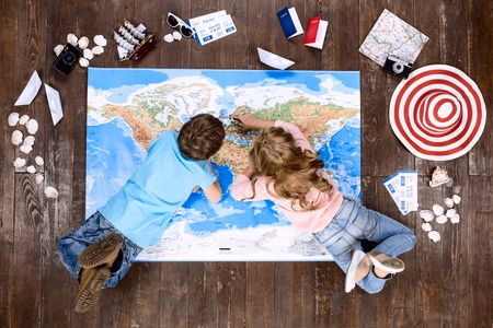 Happy children. Top view creative photo of little boy and girl on vintage brown wooden floor. Children lying on world map near travel thingsの写真素材