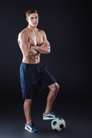 Attractive and muscular football player. Studio shot of young shirtless sportsman on black background. Man with football ball looking at cameraの写真素材