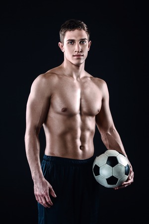 Attractive and muscular football player. Studio shot of young shirtless sportsman on black background. Man with football ball looking at cameraの写真素材