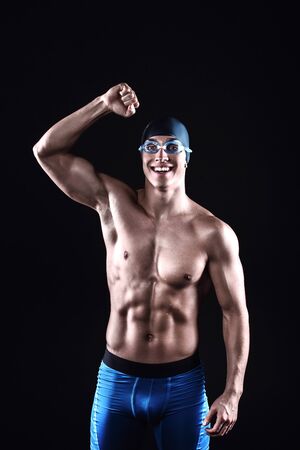 Attractive and muscular swimmer. Studio shot of young shirtless sportsman on black background. Happy man with glasses showing victoryの写真素材