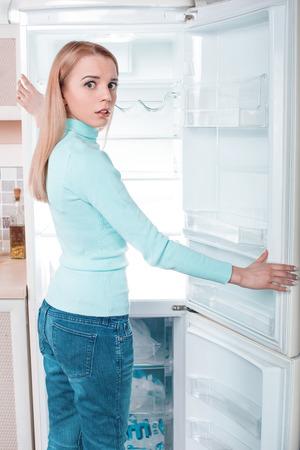 What's for dinner? Pretty blonde standing near open empty fridge. Back view photo of thoughtful young woman. She looking at cameraの写真素材