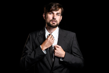Stylish young businessman on black background. Businessman looking at camera and correcting tieの写真素材