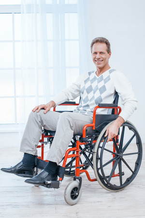 Adult man in wheelchair. White interior with big window. Man smiling and looking at cameraの写真素材