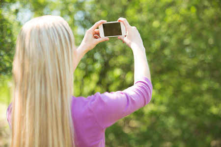 Nice looking young woman outdoors. Woman making selfie photo with mobile phone. Beautiful green park as a backgroundの写真素材