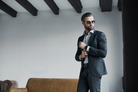 Young bearded stylish businessman leader indoors at office wearing sunglasses walking fixing sleeves with watch looking aside seriousの写真素材
