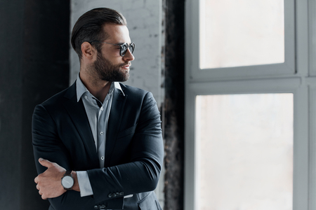 Young bearded stylish businessman leader indoors at office wearing sunglasses walking fixing sleeves with watch looking aside seriousの写真素材