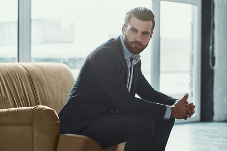 Young bearded stylish businessman leader indoors at office sitting looking aside holding sunglasses thoughtfulの写真素材