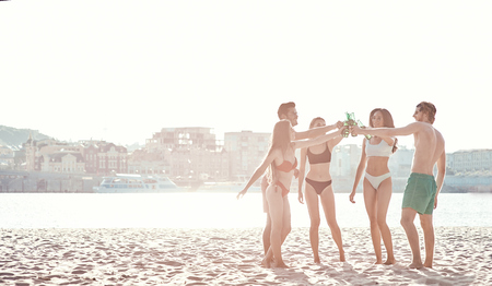 Enjoying carefree time with friends. Cheerful young people spending nice time together while sitting on the beach and drinking beerの写真素材