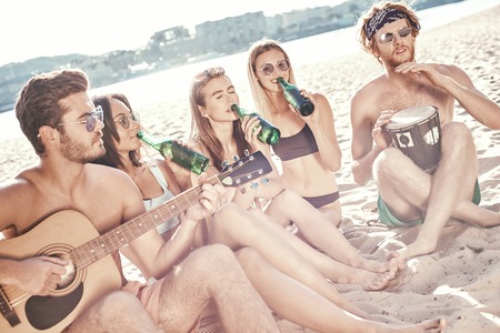 Group of young female friends sitting on beach on sunbeds,singing and playing guitar.Joying in sunset.の写真素材