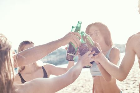 Enjoying carefree time with friends. Cheerful young people spending nice time together while sitting on the beach and drinking beerの写真素材