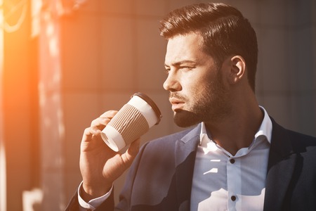 Close-up of young man holding coffee to take away at early morning in sunny park, sunlight, blurred backgroundの写真素材