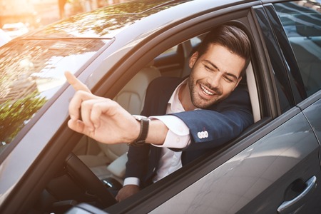 Portrait of businessman driving in car and smiling on his morning commute to work. Young man his finger pointing towards the camera.の写真素材