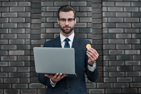 Photo of handsome rich businessman in suit showing golden bitcoin and laptop focused on cameraの写真素材
