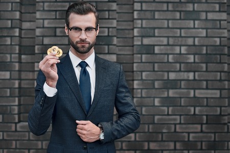 Photo of handsome rich businessman in suit showing golden bitcoin focused on cameraの写真素材