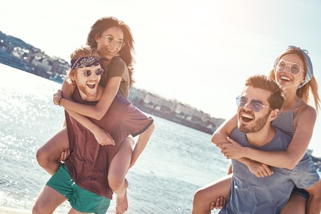 Outdoors photo of happy boyfriends piggybacking their girlfriends at sunset on beachの写真素材