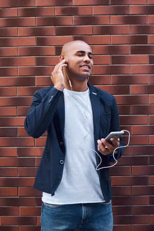 Music lover. Young guy wearing earphones standing on wall listening to music on smartphone closed eyes smiling cheerfulの写真素材