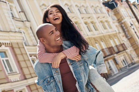 Romantic Relationship. Young diverse couple walking on the city street boyfriend carrying girlfriend on back laughing happyの写真素材