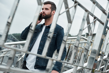 Outdoors leisure. Young stylish man walking down the stairs talking on smartphone smiling joyful view through fence close-upの写真素材