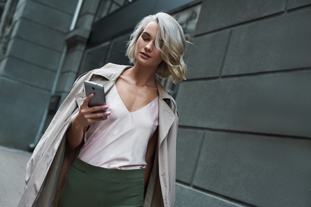 Fashion. Young stylish woman walking on the city street browsing internet on smartphone concentratedの写真素材