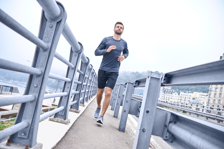 Always in great shape. Full length of young man in sports clothing exercising while jogging on the bridge outdoorsの写真素材