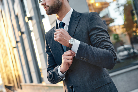 Cropped profile portrait of a successful young bearded guy in suit and glasses. So stylish and nerdy. Outdoors on a sunny street, fixing his cuffsの写真素材