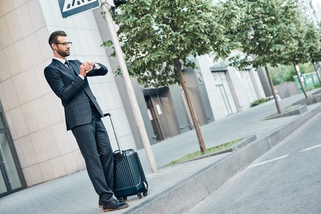 Handsome bearded businessman in classic suit with suitcase is waiting for a taxi, side viewの写真素材