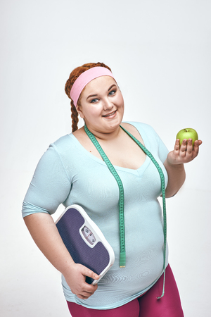 Funny picture of amusing, red haired, chubby woman on white background. Woman holding an apple and scales.の写真素材