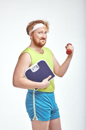 Funny picture of red haired, bearded, plump man on white background. Man holding a dumbbell and scalesの写真素材