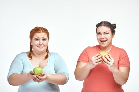 Funny picture of amusing chubby women on white background. One woman holding a sandwich, the other holding an apple.の写真素材