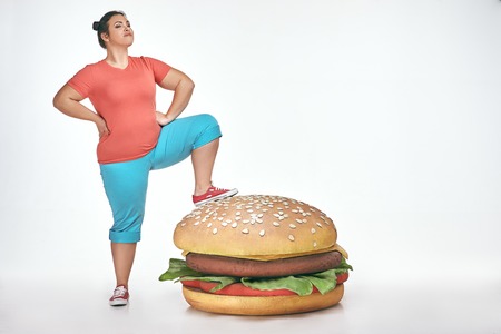 Funny picture of brunette, bearded, plump woman on white background. Woman wearing sportswear. She put her leg to a huge sandwichの写真素材