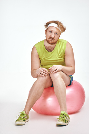 Funny picture of red haired, bearded, plump man on white background. Man wearing sportswear. Man sitting on a ball.の写真素材