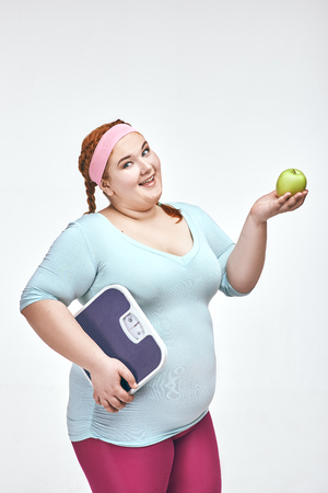 Funny picture of amusing, red haired, chubby woman on white background. Woman holding an apple and scales.の写真素材