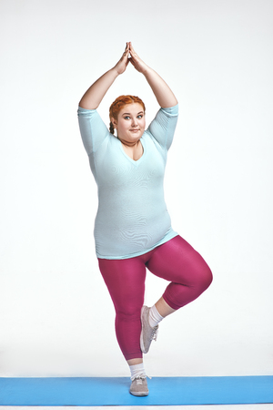 Funny picture of amusing, red haired, chubby woman on white background. Woman trains on the mat.の写真素材