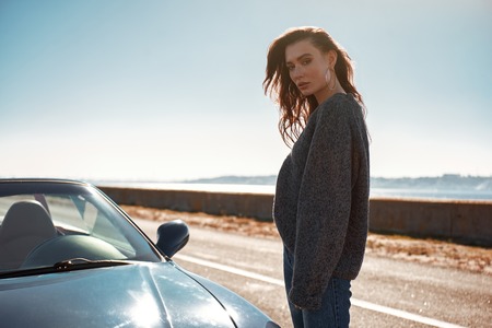 Young woman standing near roofless car outdoors on sunny day near the roadの写真素材