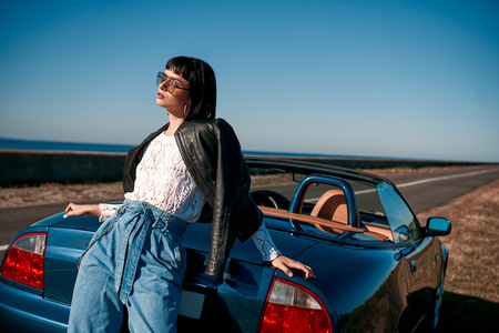 Young woman near roofless car and smiling outdoors on sunny day near the empty road. No people, sportive blue carの写真素材