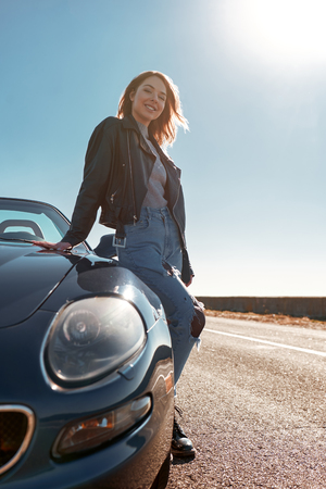 Young woman near roofless car and smiling outdoors on sunny day near the empty road. No people, sportive black carの写真素材
