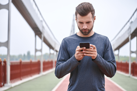 Male runner in white windbreaker typing something on his mobile smart phone standing at sunset on beautiful road, black man holding smart phone resting typing something, fit man with mobile smartphoneの写真素材