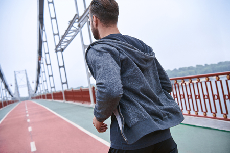 Morning workout. Close up of young man in sports clothing exercising while jogging on the bridge outdoorsの写真素材