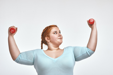 Funny red haired, chubby woman is smiling and holding dumbbellsの写真素材