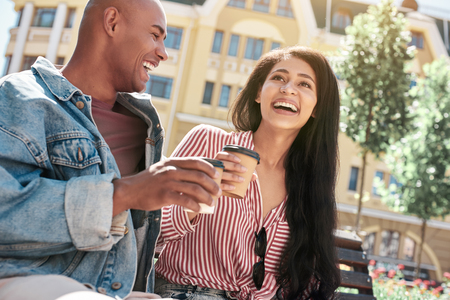 Romantic Relationship. Young diverse couple sitting on bench on the city street holding cups drinking hot coffee talking laughing cheerful close-upの写真素材