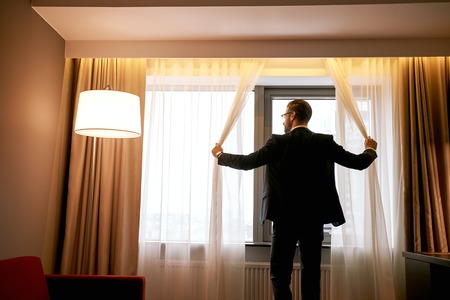 Businessman in dark hotel room opens curtains on window to the morning lightの写真素材