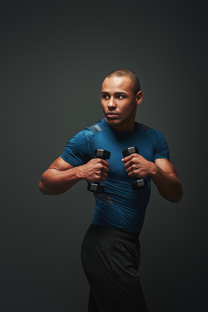 Training to become the best. Dark skinned sportsman standing with dumbbells over dark background, looking awayの写真素材