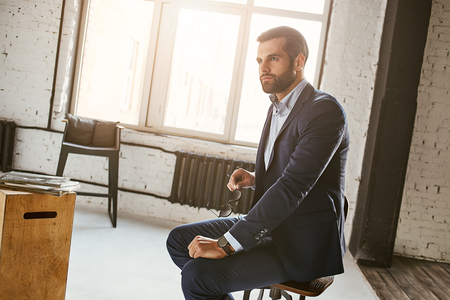 Thinking about business...Portrait of attractive businessman wearing stylish suit sitting in armchair and looking awayの写真素材