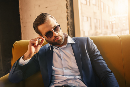 Sexy bearded businessman in sunglasses dressed in very fashionable and stylish suit is sitting at office and looking awayの写真素材