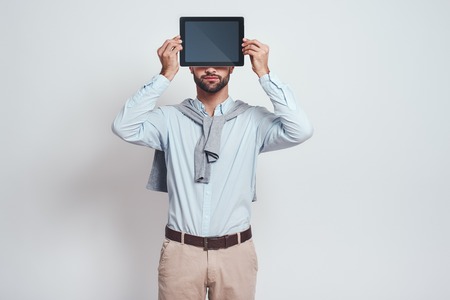Bearded man in casual clothes is holding his digital tablet in front of his head while standing against grey background. Digital concept.の写真素材
