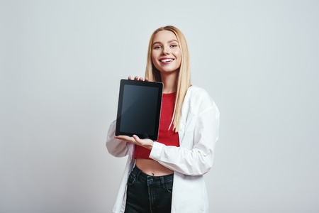 Excited young blonde woman holding and showing the screen of her modern touchpad. Digital concept