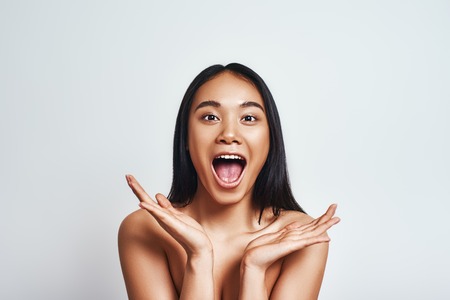 Surprised young asian woman keeping palms opened and looking at camera while standing against grey background. Positive emotionsの写真素材