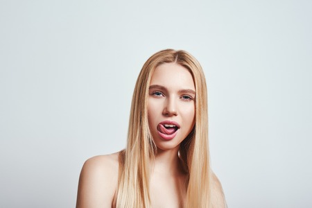 Bad girl. Close-up portrait of playful young woman with long hair looking at camera and sticking out tongue while standing against grey background